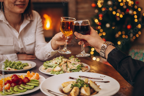 hands with glasses clinking against the background of Christmas tree lights and bonfires from a home fireplace over a table with delicious dishes