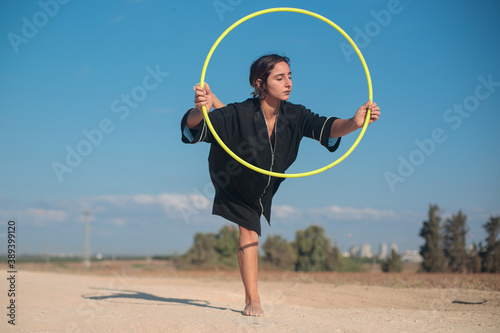 Female flow artist dancing with a Hula hoop outdoors, with one leg on the ground. Wide shot in natural morning sunlight. 