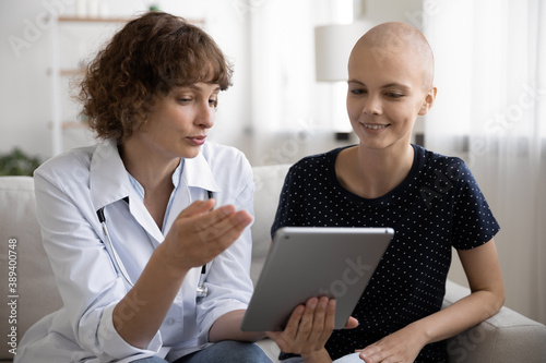 Close up doctor consulting hairless woman about medical checkup result, holding computer tablet, sitting on couch in hospital, nurse explaining treatment plan, young female struggling with cancer