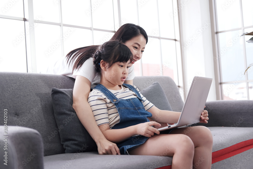 Smiling Asian mother and little asian girl child is studying and doing homework with laptop on sofa. Homeschool and online learning with technology for quarantine during Coronavirus epidemic concept. Smiling Asian mother and little asian girl child is studying and doing homework with laptop on sofa. Homeschool and online learning with technology for quarantine during Coronavirus epidemic concept.