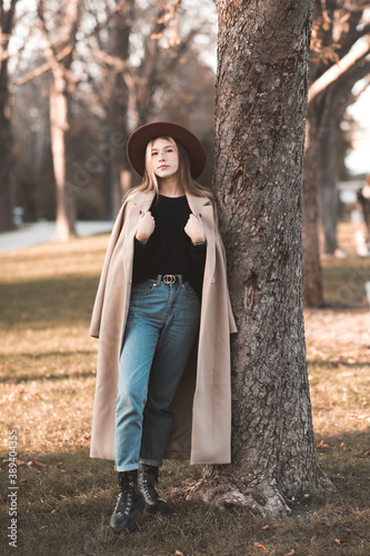 Stylish teenage girl 14-15 year old wearing trendy winter coat and hat posing in park lean on tree outdoors. Looking at camera. Teenagerhood.