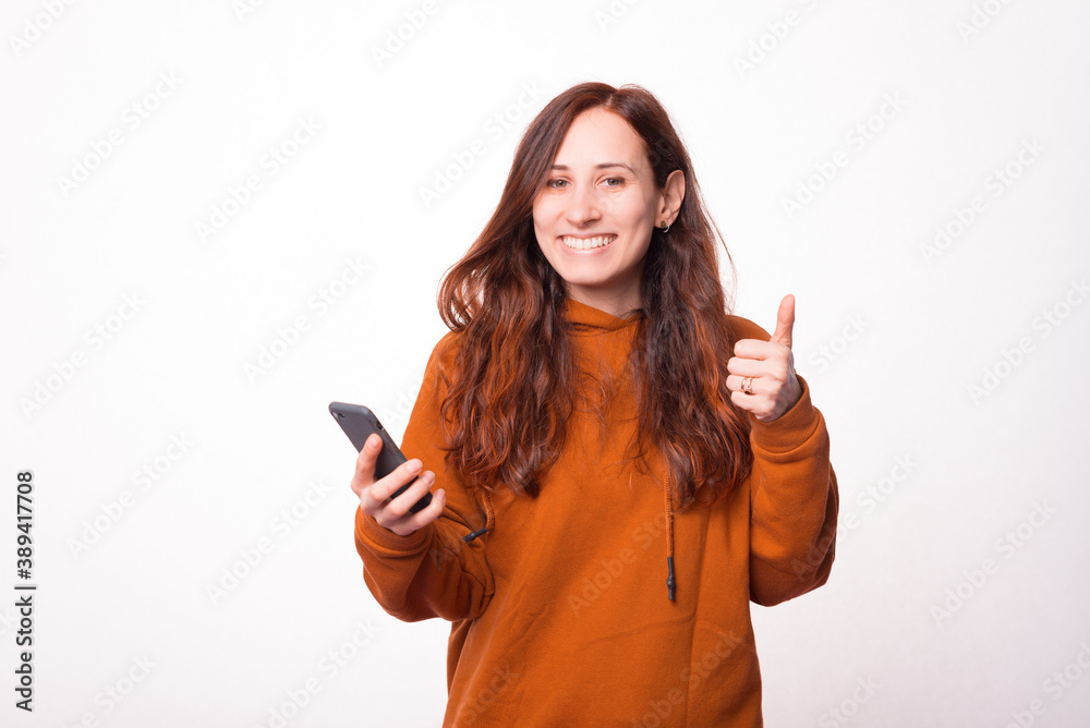Happy young woman is holding a phone and showing a thumb up is smiling at the camera . Happy young woman is holding a phone and showing a thumb up is smiling at the camera .