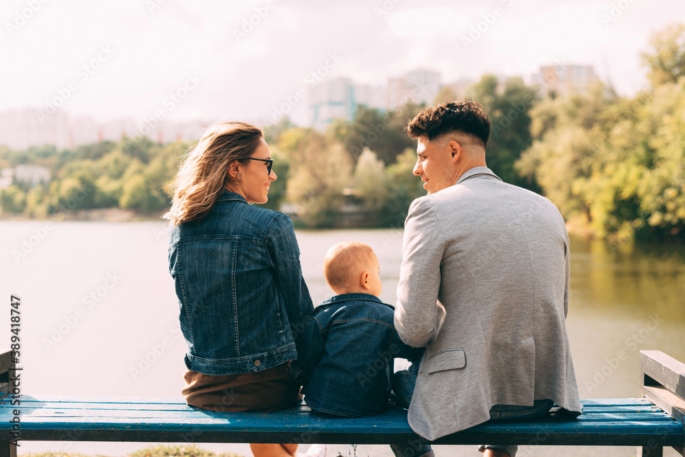 Picture of a young family in park looking at each other near a lake on a bench .