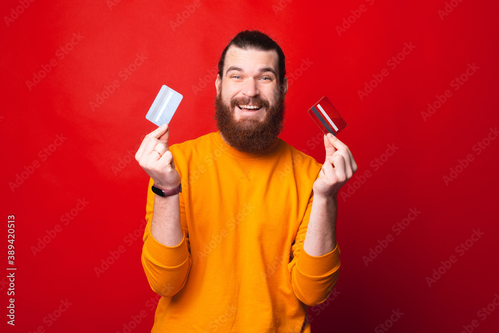 Excited young bearded man is looking at the camera and holding two credit cards . Excited young bearded man is looking at the camera and holding two credit cards .