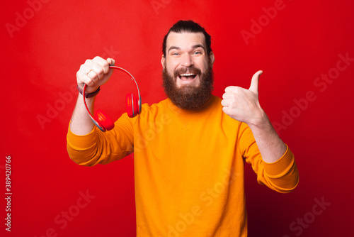 Photo of a bearded young man holding headphones and showing a thumb up is smiling at the camera .