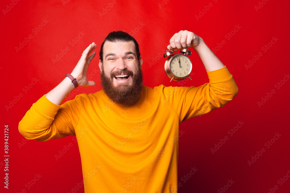 A bearded man is holding a clock looking at the camera is smiling . A bearded man is holding a clock looking at the camera is smiling .