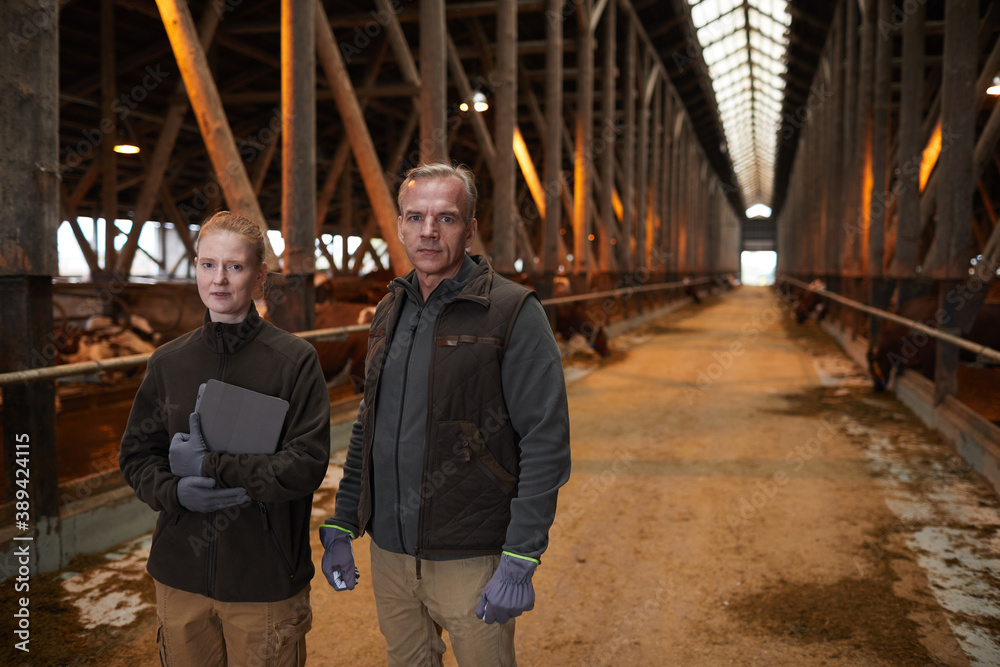 Waist up portrait of father and daughter standing in barn and looking at camera while working at family farm, copy space