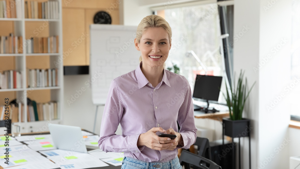 Portrait of smiling young european blonde businesswoman standing in office, holding smartphone in hands, successful female employee manager using modern technology gadget for effective communication. Portrait of smiling young european blonde businesswoman standing in office, holding smartphone in hands, successful female employee manager using modern technology gadget for effective communication.