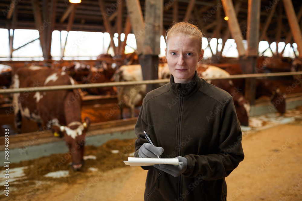 Waist up portrait of young female worker holding clipboard and looking at camera while standing in livestock shed at family farm, copy space Waist up portrait of young female worker holding clipboard and looking at camera while standing in livestock shed at family farm, copy space