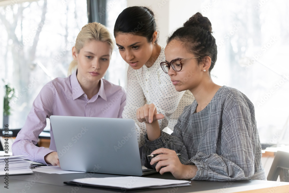 Skilled young indian mixed race businesswoman helping concentrated multiracial female interns with corporate computer software, focused diverse colleagues working on online project presentation. Skilled young indian mixed race businesswoman helping concentrated multiracial female interns with corporate computer software, focused diverse colleagues working on online project presentation.