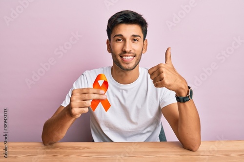 Young handsome man holding awareness orange ribbon smiling happy and positive, thumb up doing excellent and approval sign photo