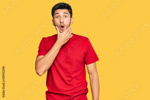 Young handsome man wearing casual red tshirt looking fascinated with disbelief, surprise and amazed expression with hands on chin