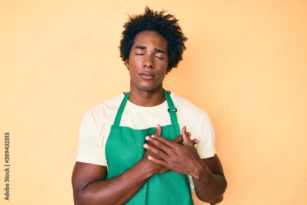 Handsome african american man with afro hair wearing waiter apron smiling with hands on chest with closed eyes and grateful gesture on face. health concept. Handsome african american man with afro hair wearing waiter apron smiling with hands on chest with closed eyes and grateful gesture on face. health concept.