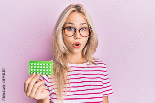 Beautiful caucasian blonde girl holding birth control pills scared and amazed with open mouth for surprise, disbelief face