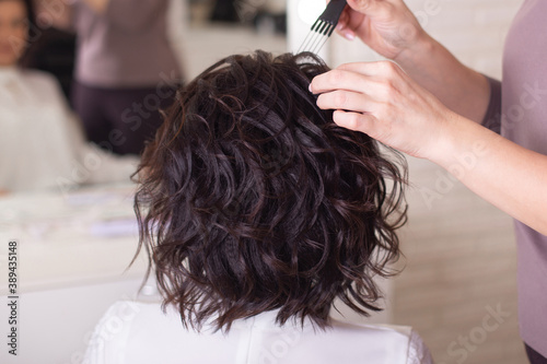 Female hands of hairstylist and back of brunette woman with short wavy hair on beauty salon background