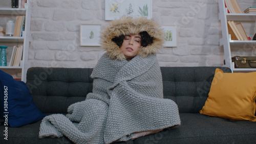 Fotografia Young sick woman sitting alone on sofa in cold home living room with blanket and jacket