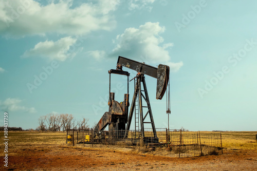 Rusty oil well pump jack  sitting in winter pasture with rough hay stubble and clay covered ground and bare trees on horizon under pretty blue sky