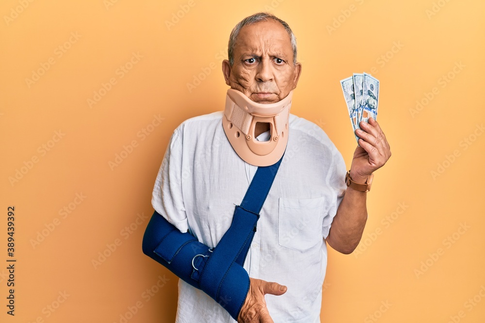 Handsome mature injured man wearing cervical collar and sling holding insurance money clueless and confused expression. doubt concept.