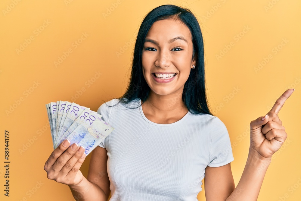 Beautiful hispanic woman holding 20 swedish krona banknotes smiling happy pointing with hand and finger to the side Beautiful hispanic woman holding 20 swedish krona banknotes smiling happy pointing with hand and finger to the side