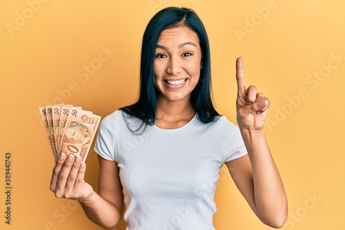 Beautiful hispanic woman holding 50 brazilian real banknotes smiling with an idea or question pointing finger with happy face  number one