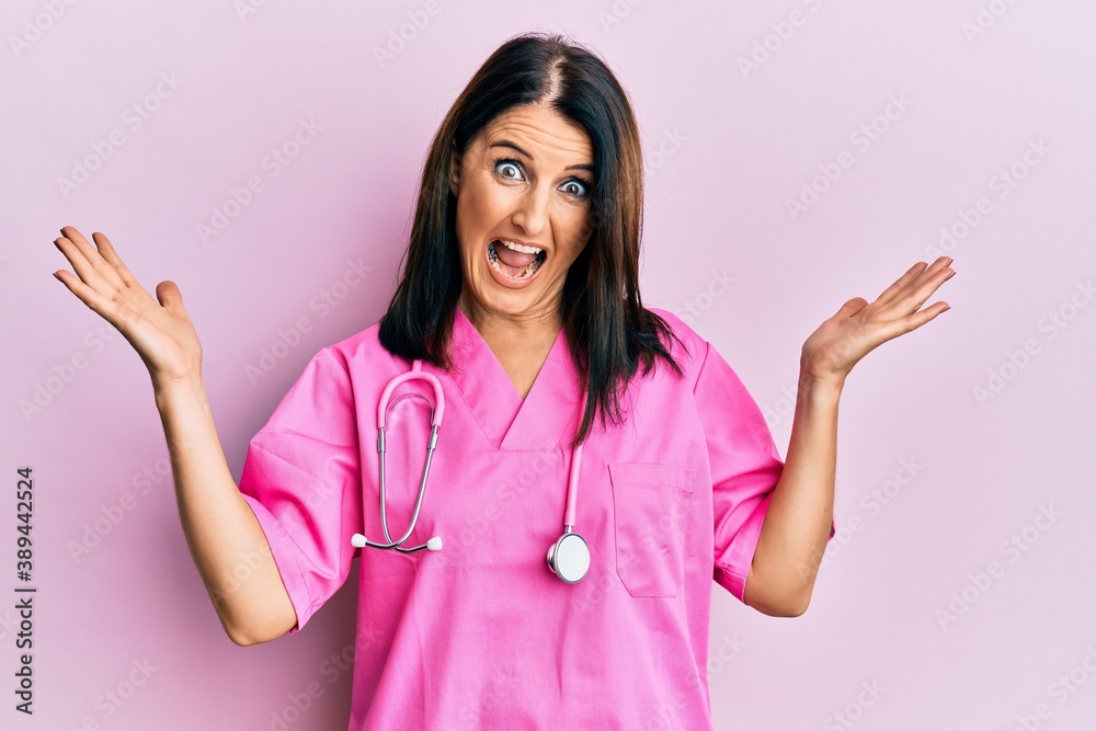 Middle age brunette woman wearing doctor uniform and stethoscope celebrating crazy and amazed for success with arms raised and open eyes screaming excited. winner concept Middle age brunette woman wearing doctor uniform and stethoscope celebrating crazy and amazed for success with arms raised and open eyes screaming excited. winner concept