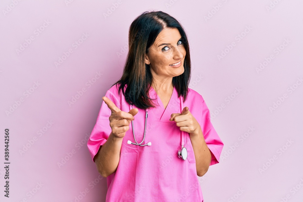Middle age brunette woman wearing doctor uniform and stethoscope pointing fingers to camera with happy and funny face. good energy and vibes. Middle age brunette woman wearing doctor uniform and stethoscope pointing fingers to camera with happy and funny face. good energy and vibes.