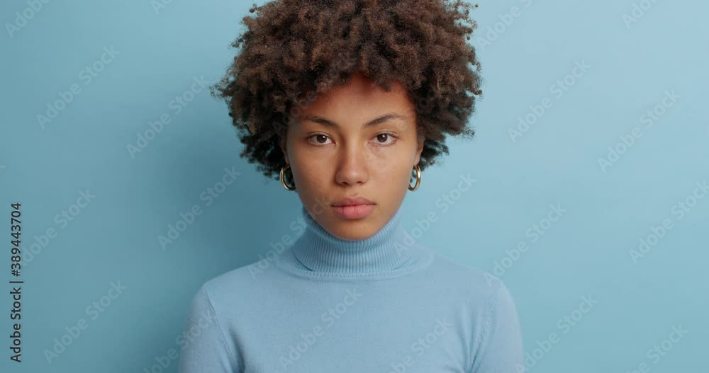 Attractive serious woman with curly hairstyle looks directly at camera dressed in casual turtleneck winks eyes has calm face expression isolated on blue wall. Assertive young Afro American female