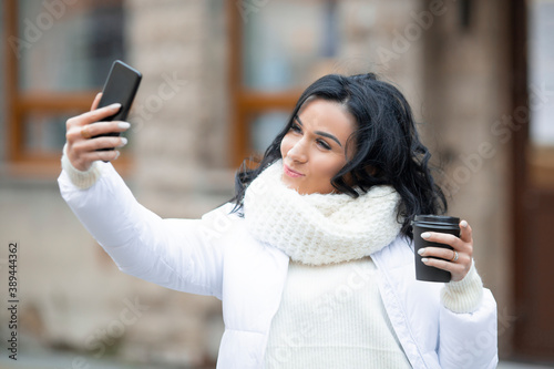 A beautiful girl with coffee in her hand makes a selfie on a mobile phone.
