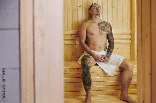 Naked fit young man in towel resting in hot sauna at wellness spa resort