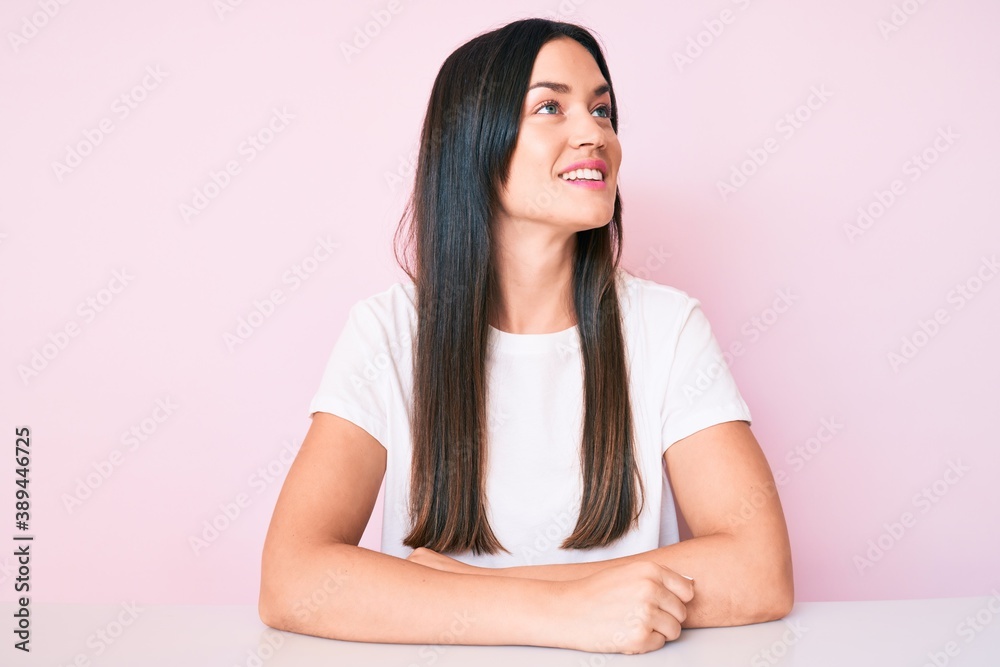 Young caucasian woman sitting at the table wearing casual white tshirt looking away to side with smile on face, natural expression. laughing confident. Young caucasian woman sitting at the table wearing casual white tshirt looking away to side with smile on face, natural expression. laughing confident.