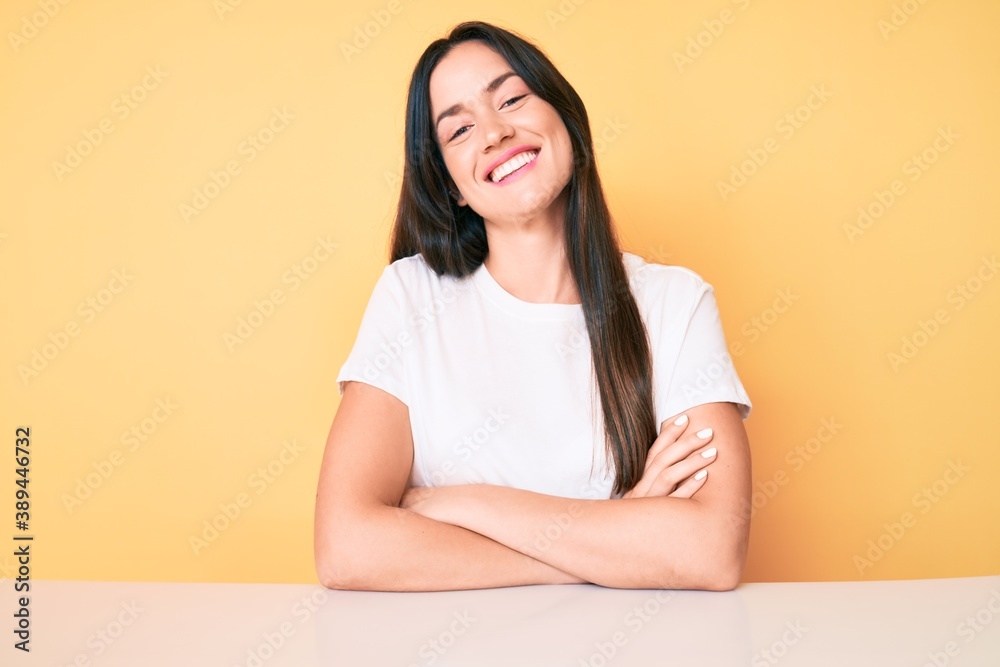Young caucasian woman sitting at the desk wearing casual white tshirt happy face smiling with crossed arms looking at the camera. positive person. Young caucasian woman sitting at the desk wearing casual white tshirt happy face smiling with crossed arms looking at the camera. positive person.