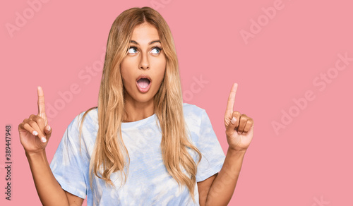 Beautiful blonde young woman wearing casual tie dye shirt amazed and surprised looking up and pointing with fingers and raised arms.