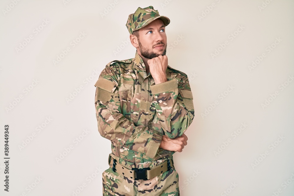 Young caucasian man wearing camouflage army uniform with hand on chin thinking about question, pensive expression. smiling and thoughtful face. doubt concept. Young caucasian man wearing camouflage army uniform with hand on chin thinking about question, pensive expression. smiling and thoughtful face. doubt concept.