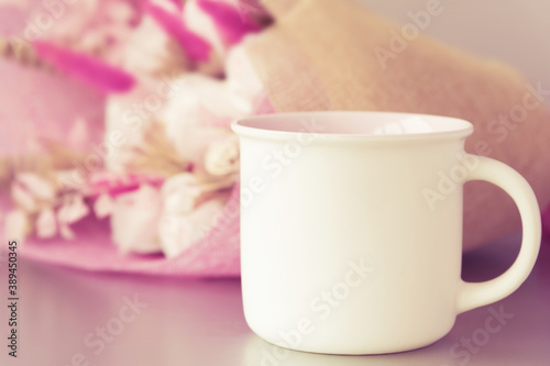 Stylish white cup with handle and space for template on side stands on light grey table against blurred bouquet extreme close view. Romantic composition.