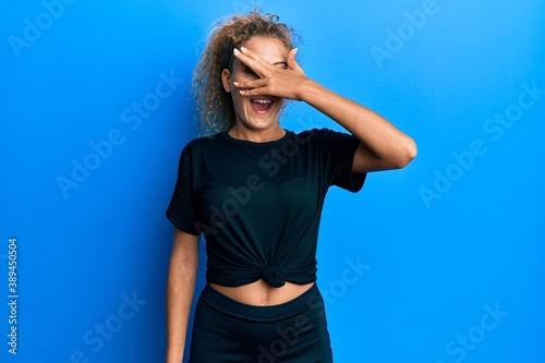 Beautiful caucasian teenager girl wearing black sportswear peeking in shock covering face and eyes with hand, looking through fingers with embarrassed expression. © Krakenimages.com