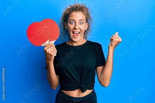 Beautiful caucasian teenager girl holding red ping pong rackets screaming proud, celebrating victory and success very excited with raised arms