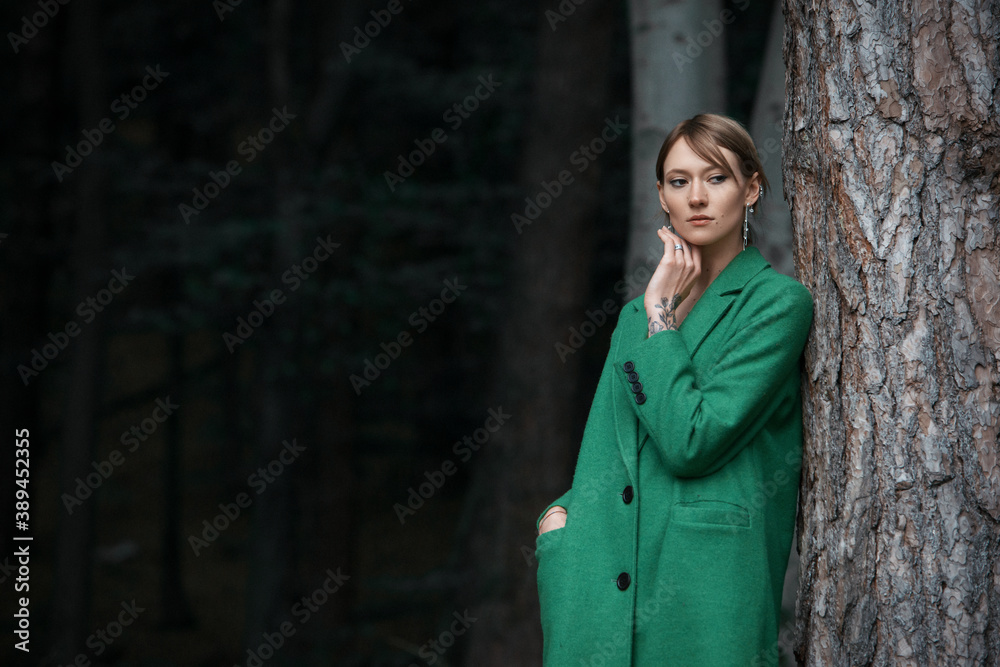 Fashionable young woman in green coat posing in a forest near the tree Fashionable young woman in green coat posing in a forest near the tree