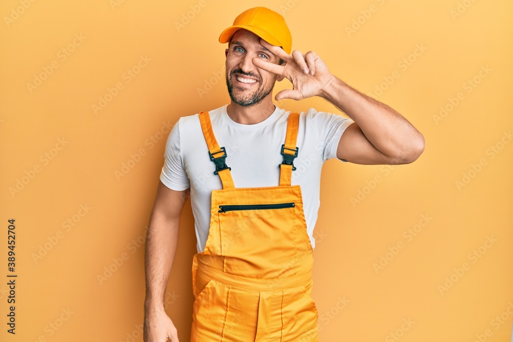 Young handsome man wearing handyman uniform over yellow background doing peace symbol with fingers over face, smiling cheerful showing victory