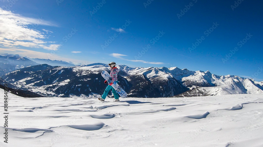 A woman walking on powder snow with her snowboard on top of Katschberg in Austria. Panoramic view on the surrounding mountains. Winter wonderland, Clear and sunny winter day. Free ride.