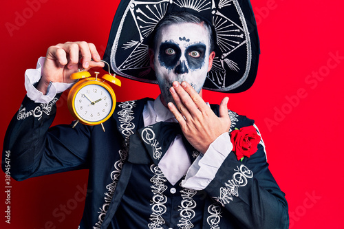 Young man wearing mexican day of the dead costume holding alarm clock covering mouth with hand, shocked and afraid for mistake. surprised expression