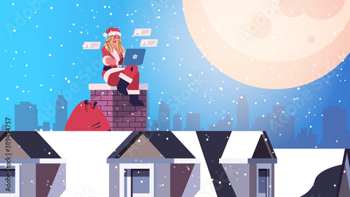 beautiful woman in santa claus costume sitting on chimney girl using laptop happy new year merry christmas holiday celebration concept cityscape backgound full length horizontal vector illustration