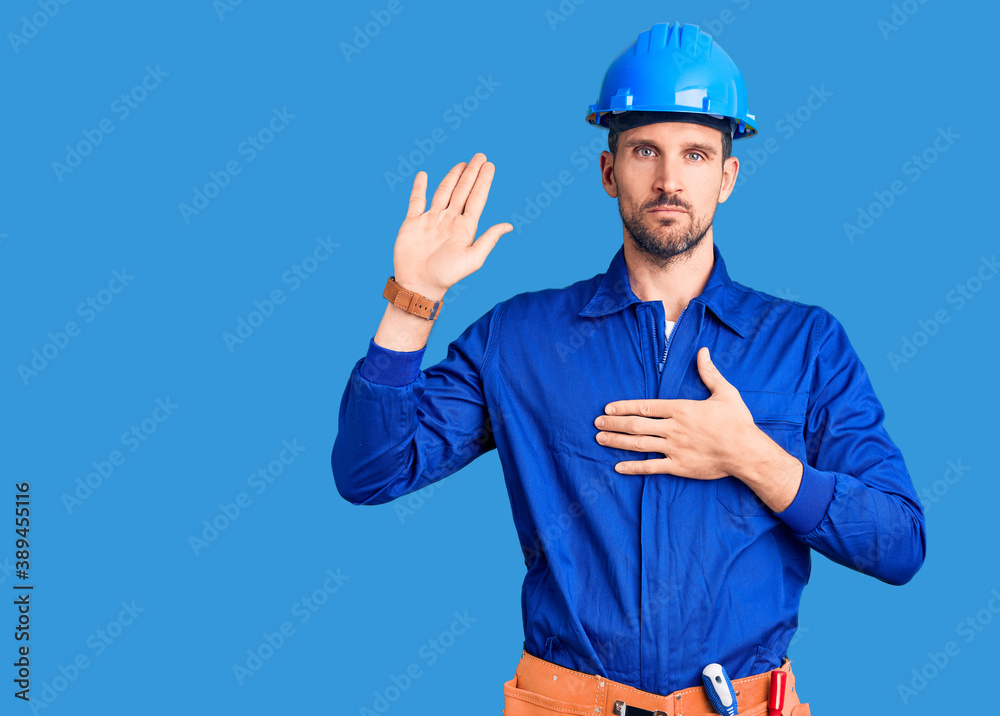 Young handsome man wearing worker uniform and hardhat covering eyes and mouth with hands, surprised and shocked. hiding emotion