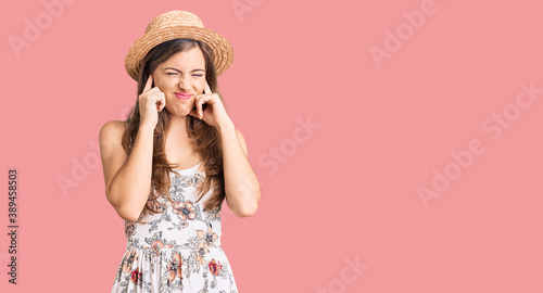 Beautiful caucasian young woman wearing summer hat covering ears with fingers with annoyed expression for the noise of loud music. deaf concept.