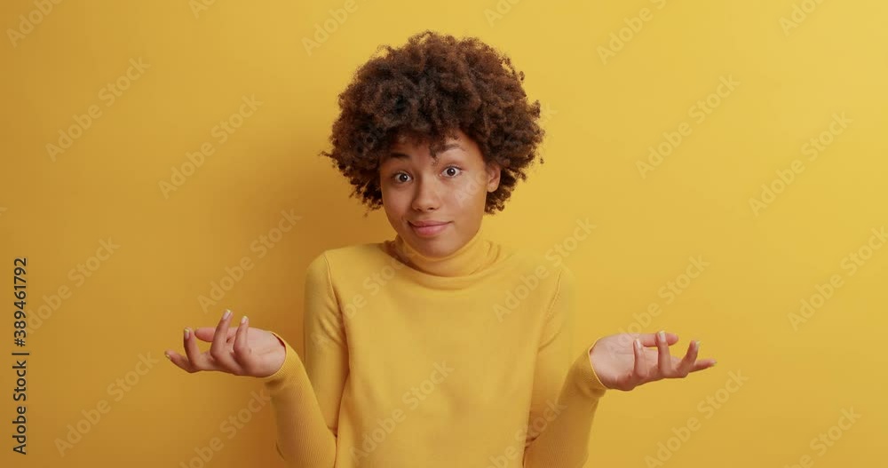 Clueless dark skinned woman throws up hands and shrugs shoulders in slow motion has doubtful expression dressed casually poses against yellow studio wall cannot help with way out makes decision