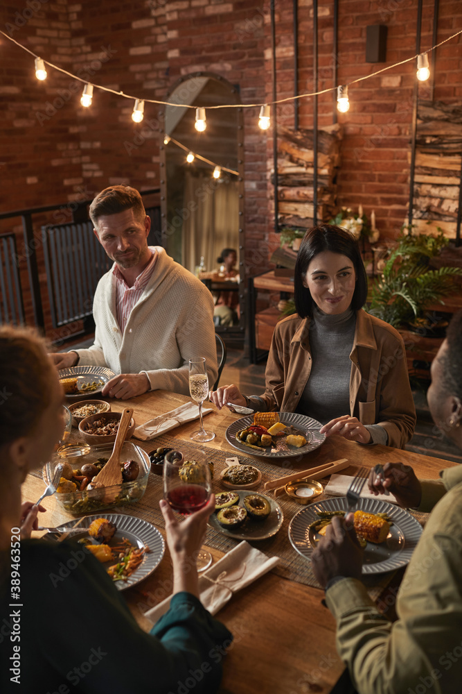 Vertical portrait of cheerful adult people sitting at dinner table while enjoying party with outdoor lighting - obrazy, fototapety, plakaty 