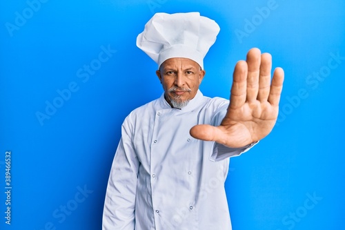 Middle age grey-haired man wearing professional cook uniform and hat doing stop sing with palm of the hand. warning expression with negative and serious gesture on the face.