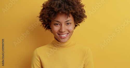 Lovely calm pleased woman with curly hair smiles broadly looks satisfied at camera being in good mood dressed in casual turtleneck expreses positive emotions isolated over yellow background.