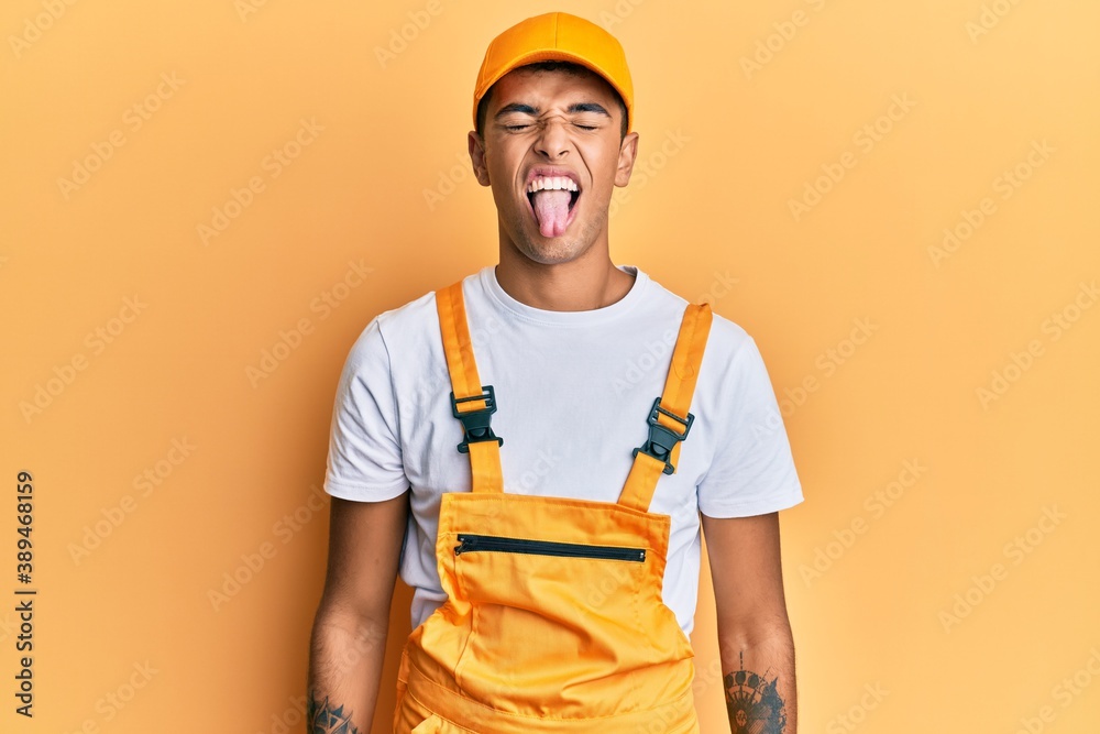 Young handsome african american man wearing handyman uniform over yellow background sticking tongue out happy with funny expression. emotion concept. Young handsome african american man wearing handyman uniform over yellow background sticking tongue out happy with funny expression. emotion concept.