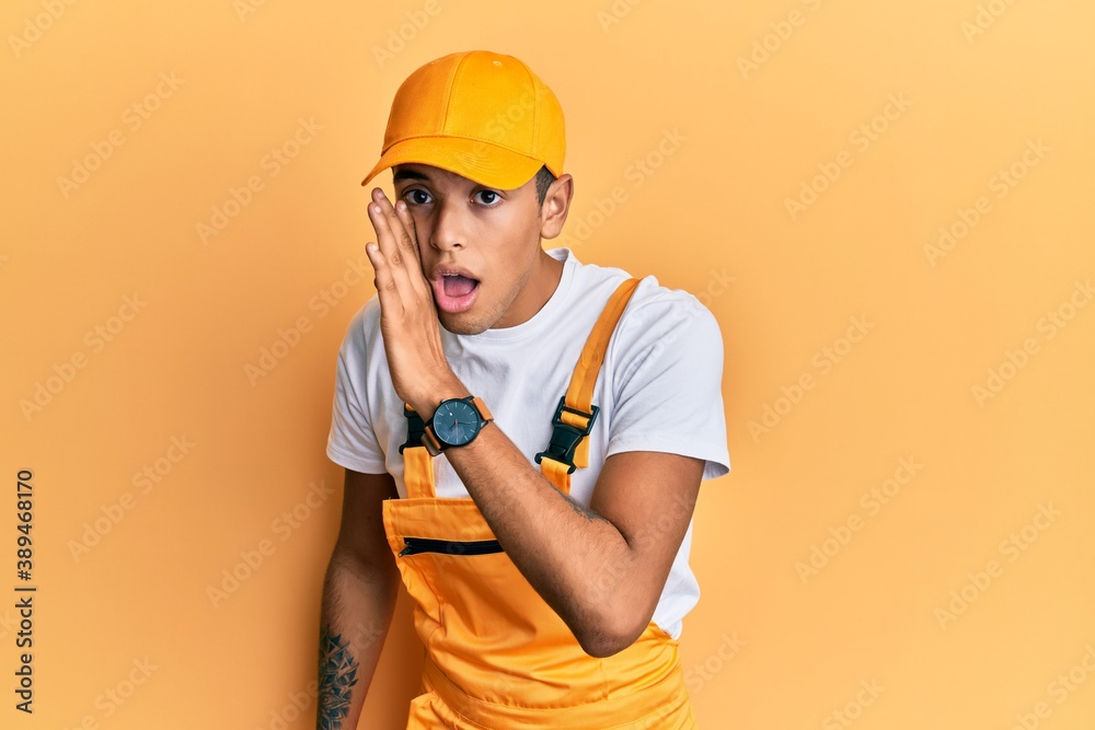 Young handsome african american man wearing handyman uniform over yellow background hand on mouth telling secret rumor, whispering malicious talk conversation Young handsome african american man wearing handyman uniform over yellow background hand on mouth telling secret rumor, whispering malicious talk conversation