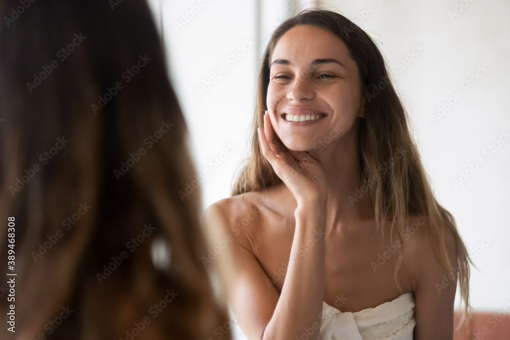 What a nice morning. Charming smiling young lady wrapped in big towel enjoying her reflection in bathroom mirror feeling happy having good mood satisfied with perfect healthy skin teeth hair condition
